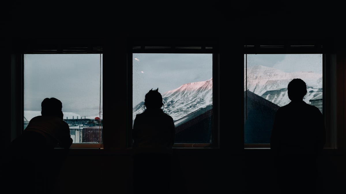 Three people at three windows of a dark room, each of them looking out at a pink-lit snow ridge