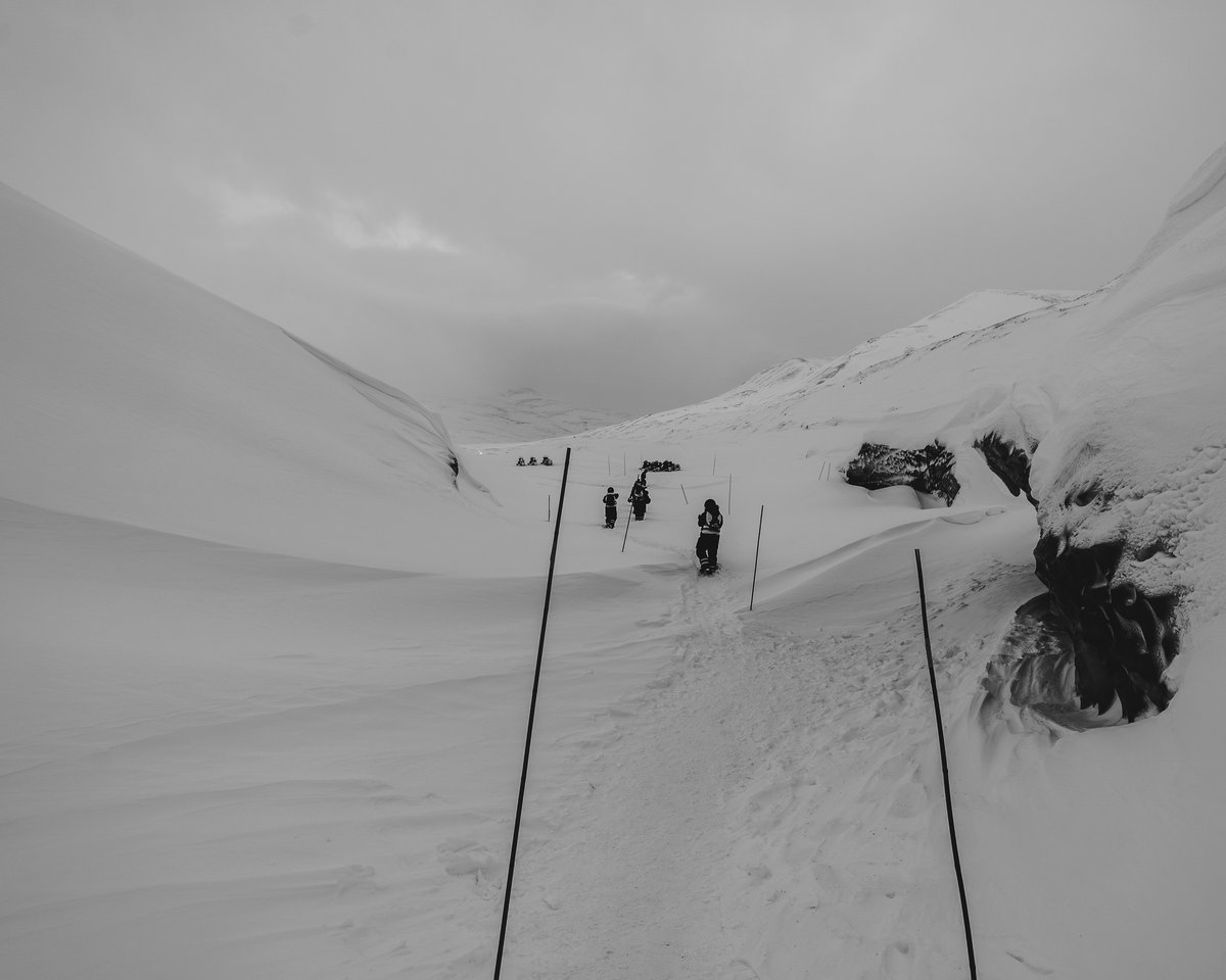 A line of people walking a marked trail up a broad snow valley, a torn blue opening in the glacier at the right