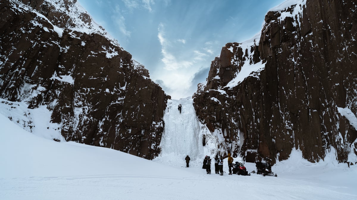 A climber partway up a frozen waterfall between two dark rock walls, the rest of the group waiting with their snowmobiles on the snow below