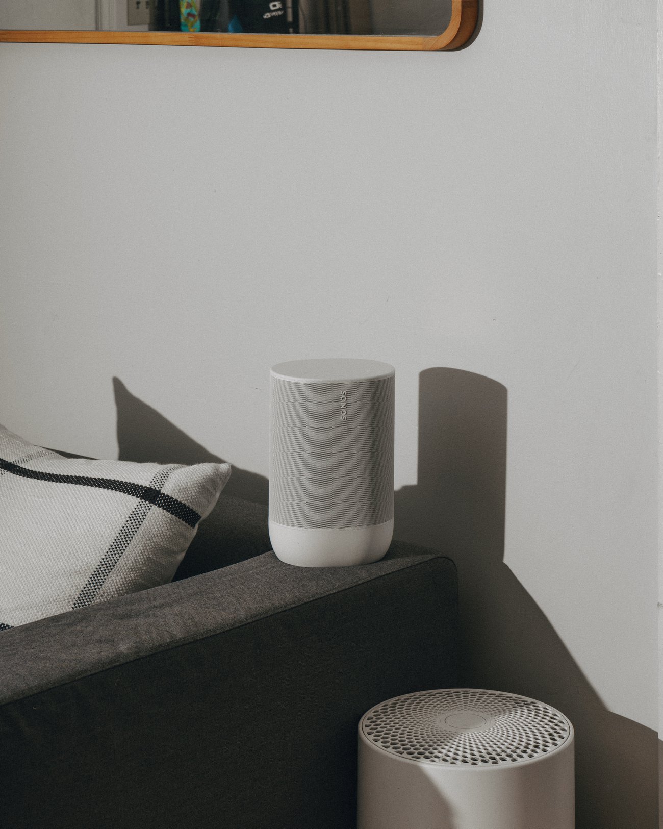 A white speaker on the arm of a dark grey sofa beside a striped cushion, casting a hard shadow on the wall, with a second speaker in the foreground