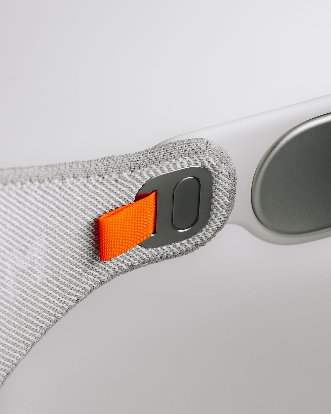 Close on the knitted head strap of a mixed-reality headset, its orange pull tab bright against the pale grey weave