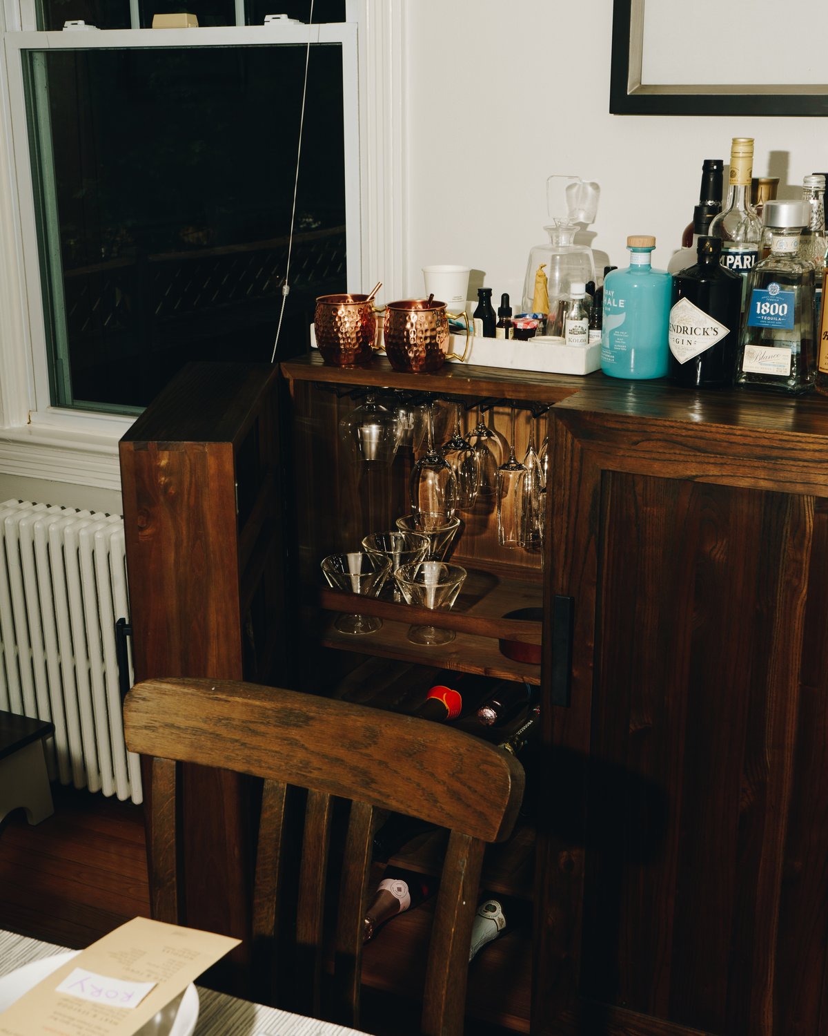 A home bar cabinet open to show hanging wine glasses and coupes, with copper mugs, gin, tequila and Campari on the top beside a laid table