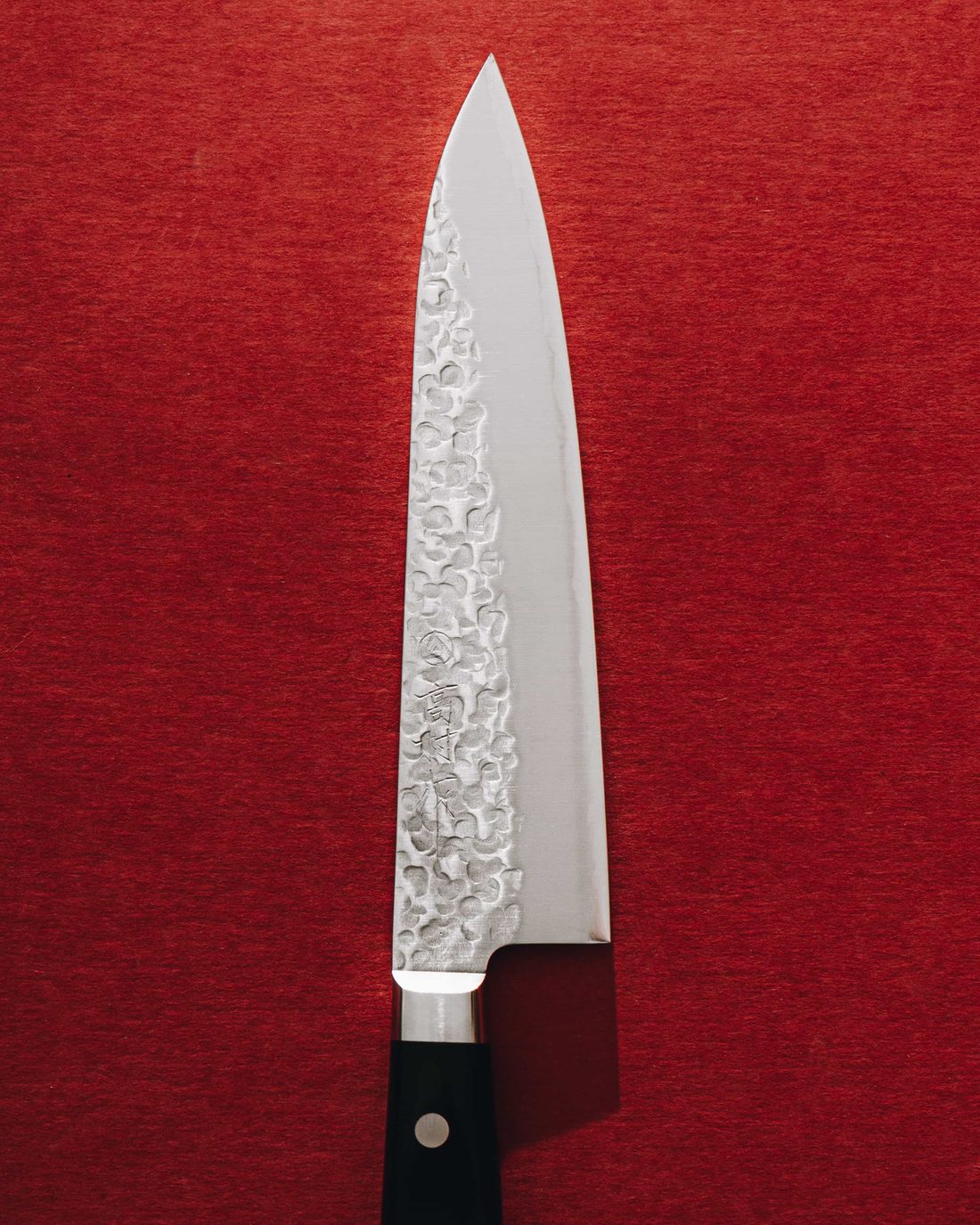 A Japanese chef's knife laid point up on red felt, hammered damascus blade and a black riveted handle