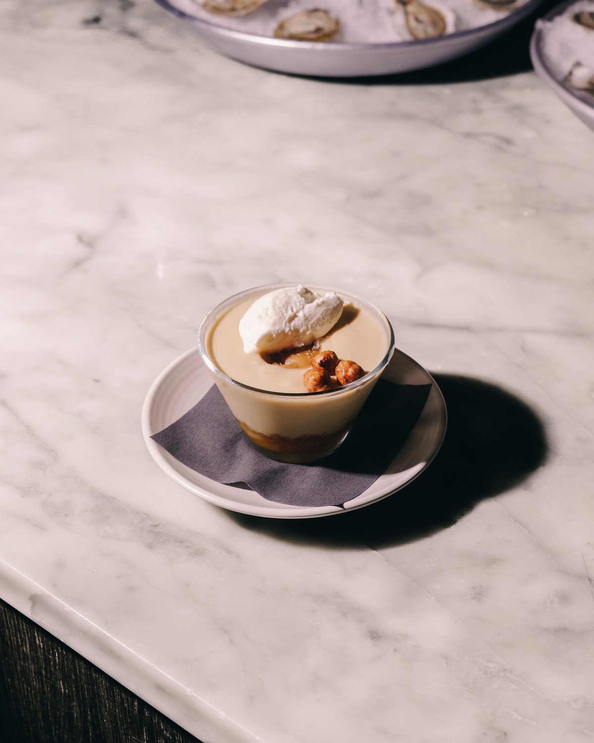 A glass of butterscotch pudding with a spoonful of cream and three glazed hazelnuts, on a saucer on a marble counter