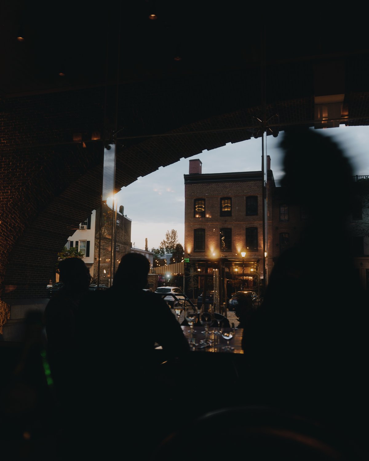 The view out of an arched restaurant window at dusk, brick warehouses and street lamps across the road, diners in silhouette at the table