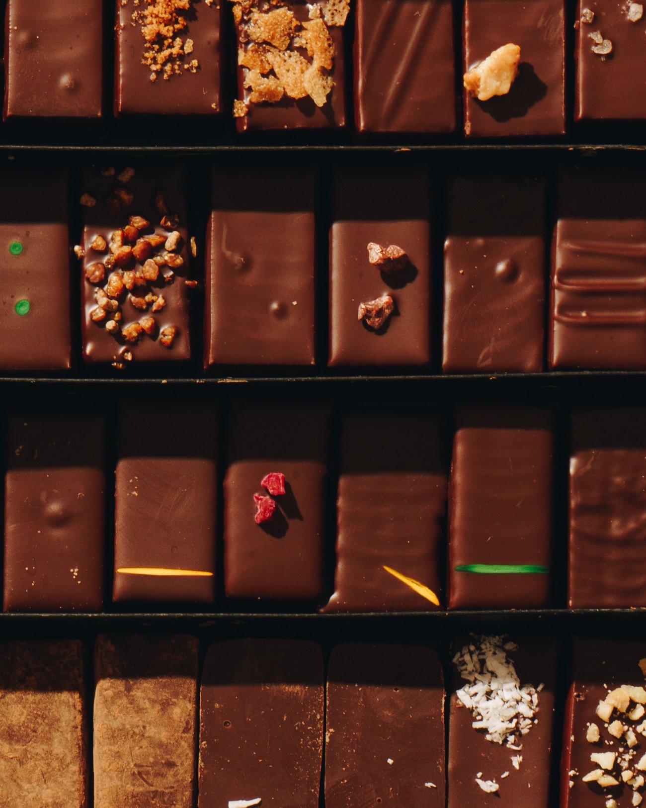 An open box of chocolates from above, four rows of rectangular pralines finished with candied peel, nuts, coconut, dried fruit and coloured lines