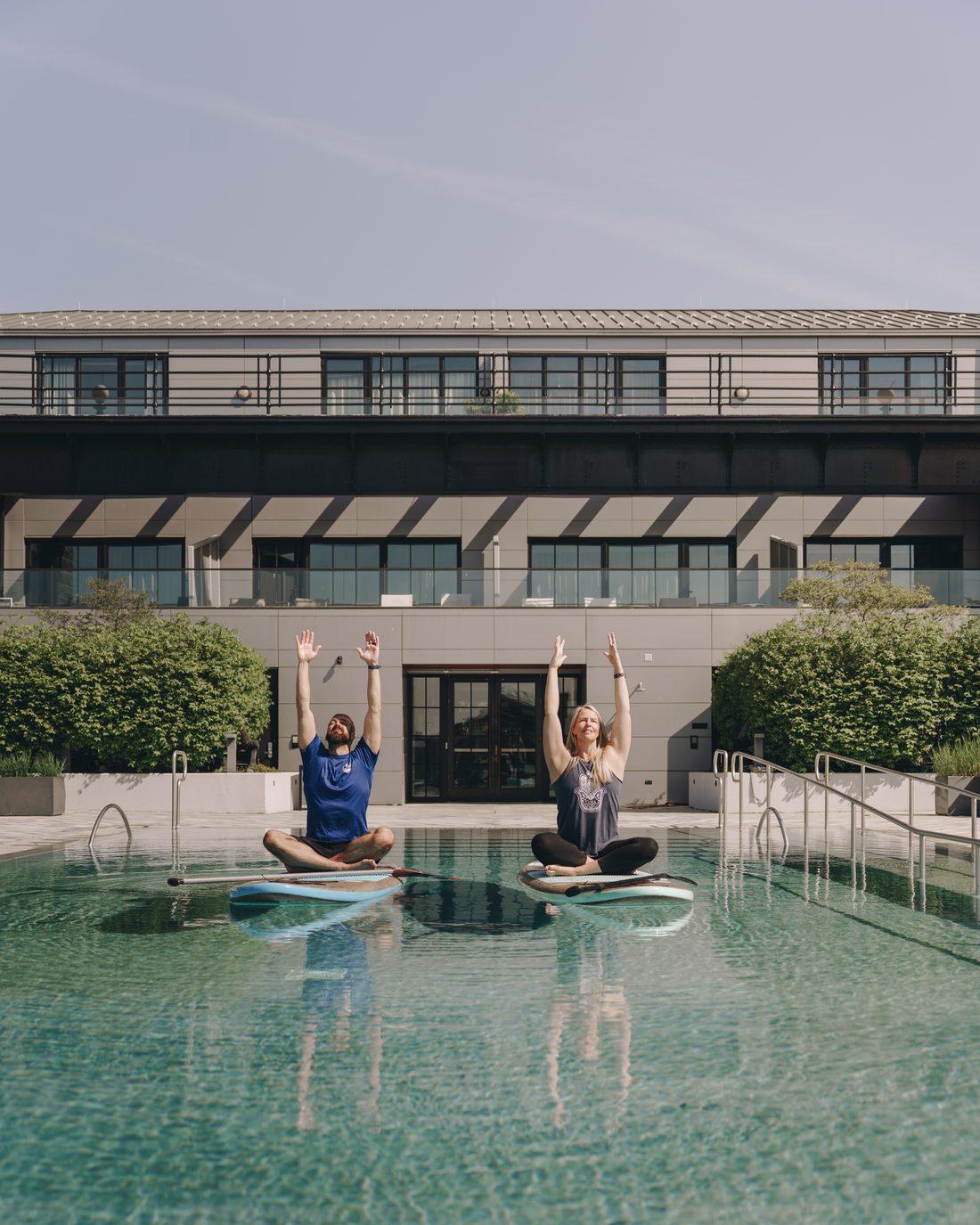 Two people sitting cross-legged on paddleboards in the pool with both arms stretched overhead
