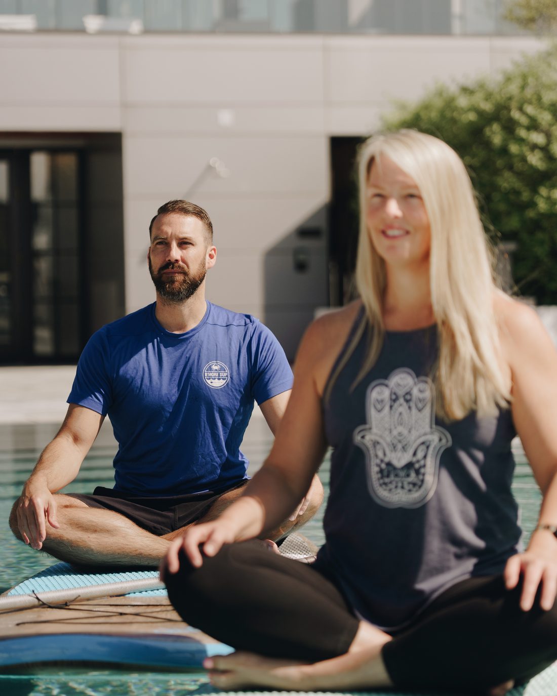 A bearded man in blue and a blonde woman in a printed tank sitting cross-legged on their boards, both laughing at the camera