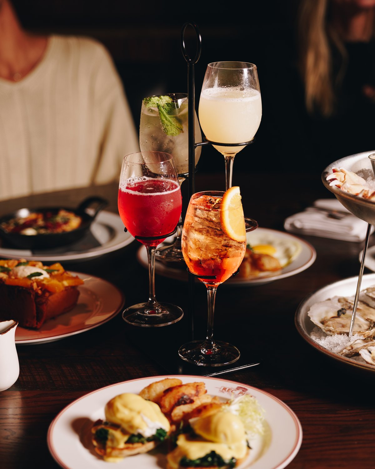 Four cocktails held in a black tiered stand, one clear with mint, one milky, one deep red and one orange over ice, with brunch plates and a bowl of oysters around them