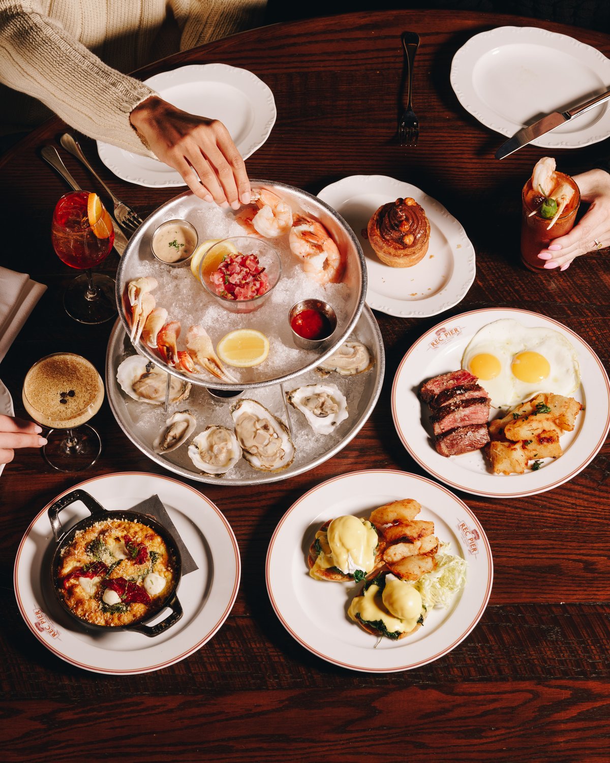 A table shot from above with a two tier seafood stand of oysters, shrimp and tuna tartare on ice, a steak and eggs plate, eggs benedict, a skillet of baked eggs and a hand reaching in for a shrimp