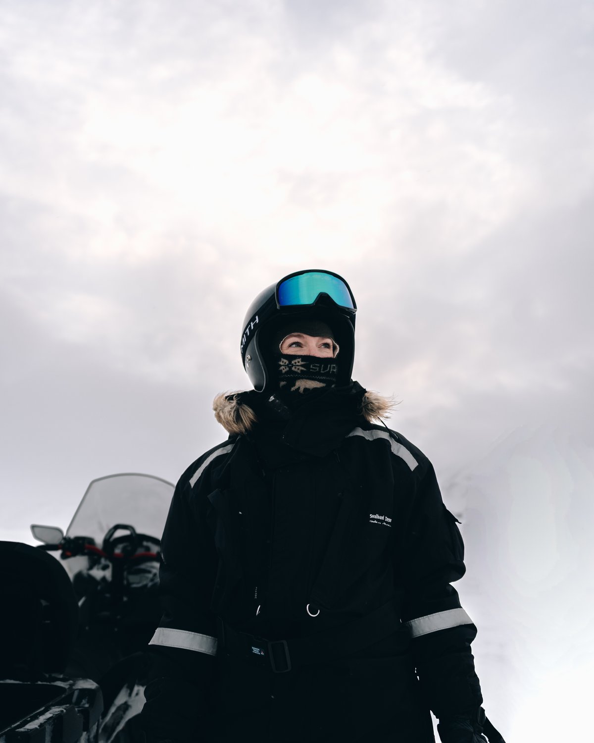 A guide in a helmet, mirrored goggles and a patterned face covering standing beside her snowmobile, looking off to one side