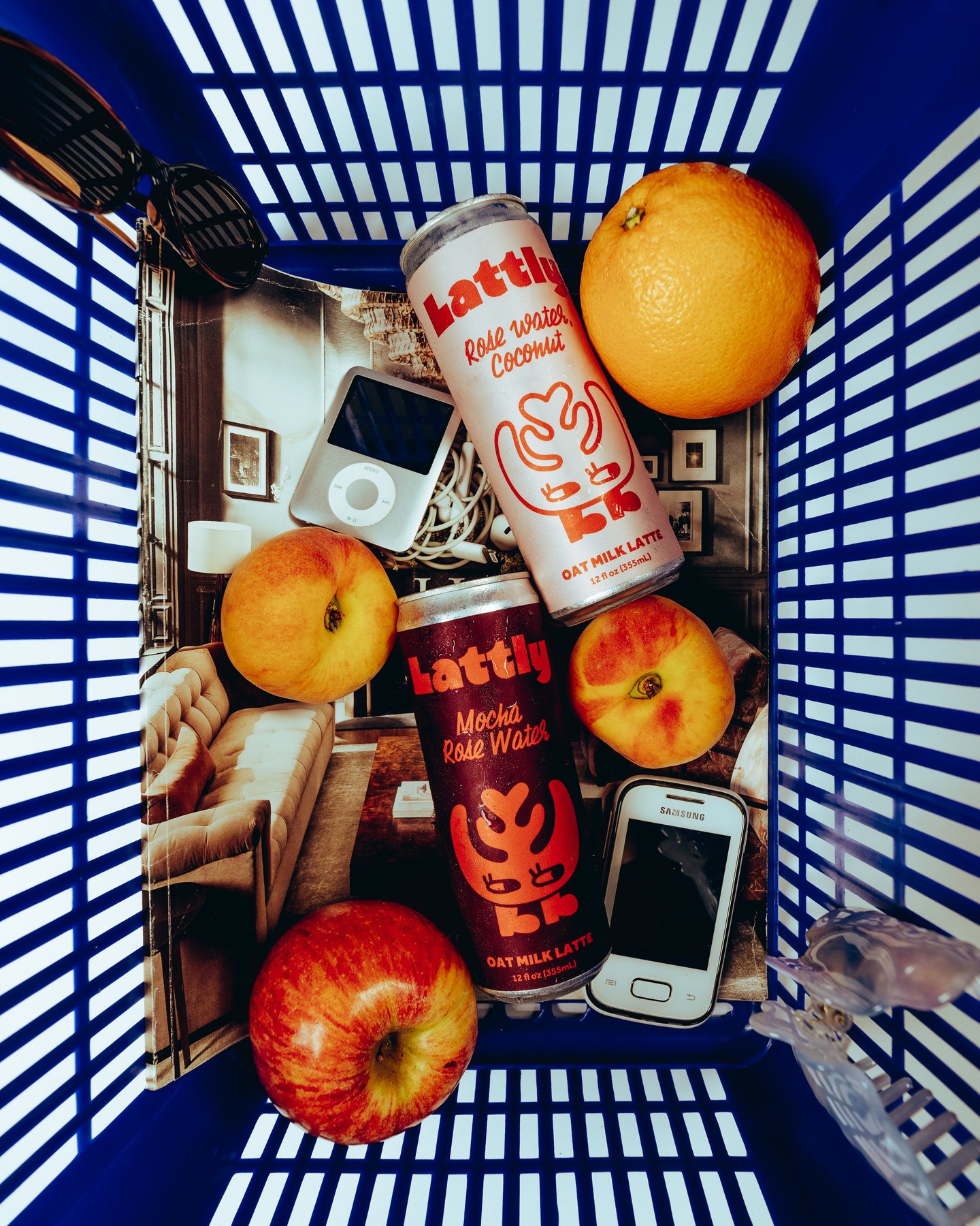 Overhead of a blue shopping basket holding oranges, an apple, two Lattly cans, a phone and a compact camera