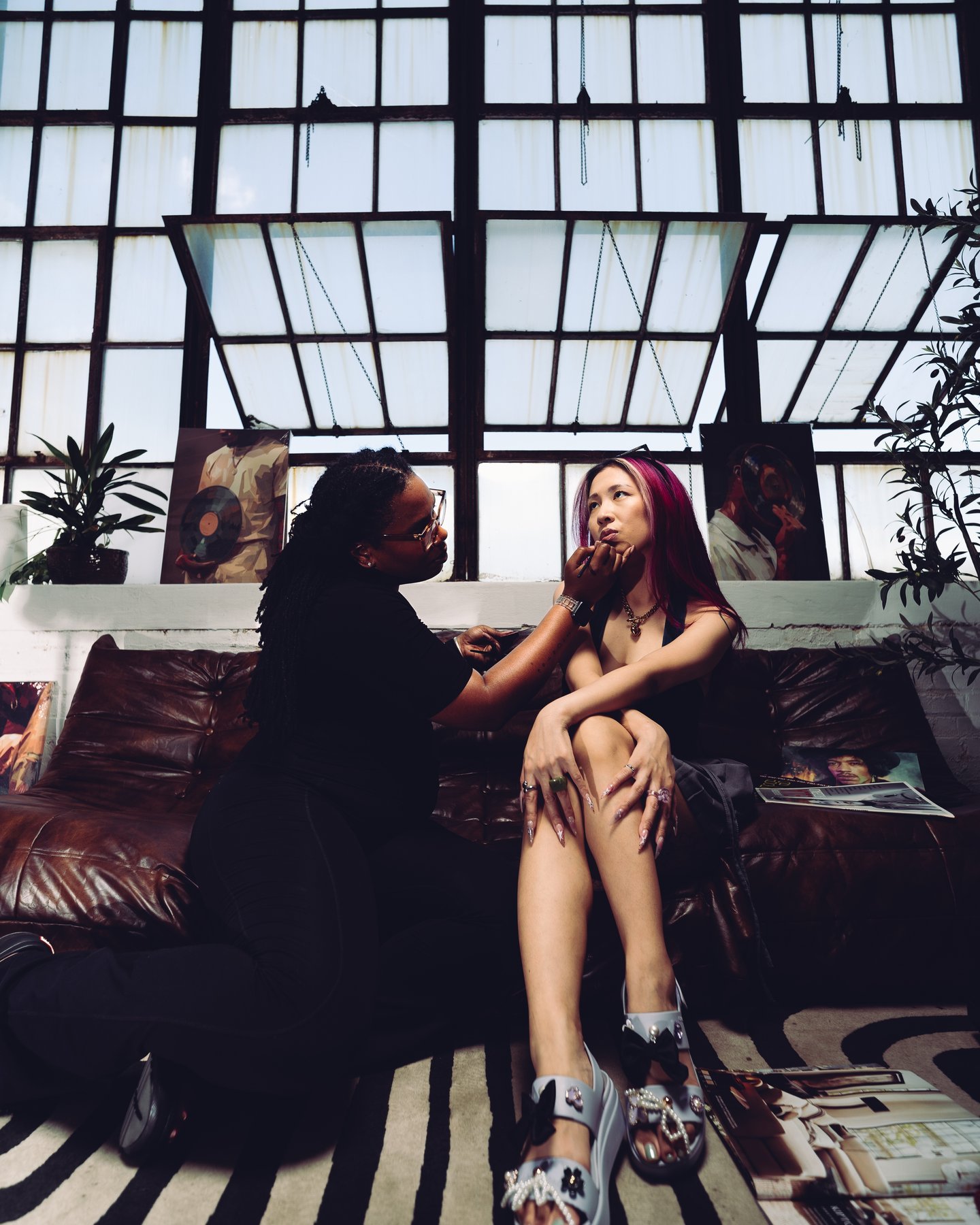 Two women on a leather sofa under a wall of loft windows, one leaning in to talk