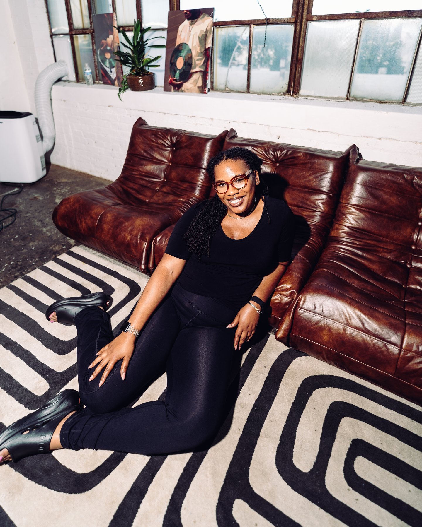 A woman in black sitting on the striped rug against the sofa, a camera in her hands