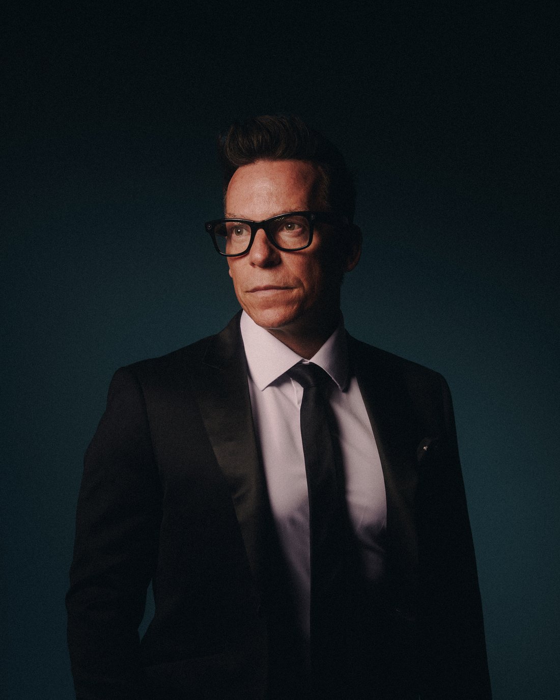 A man in a tuxedo and heavy black glasses against deep teal, looking straight down the lens