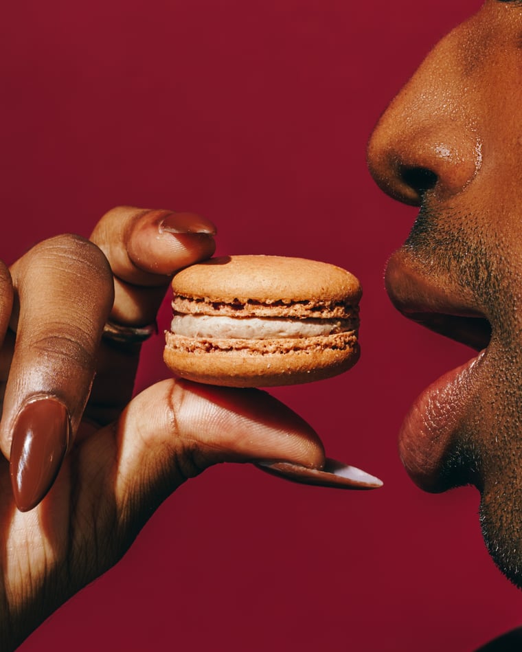 Long almond nails holding a macaron up to a pair of parted lips against deep red