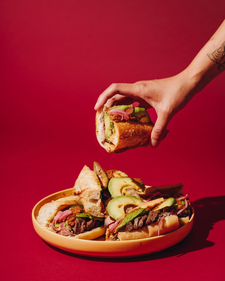 A tattooed hand lifting a loaded sandwich off a coral plate stacked with two more, on scarlet