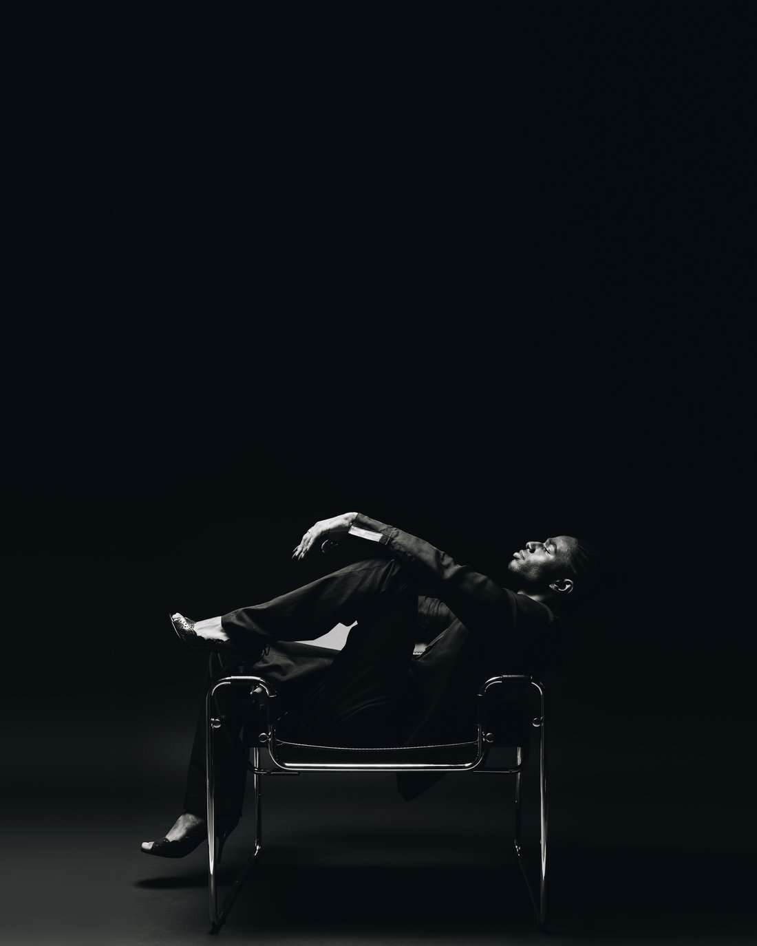 A man in a black suit thrown back across a tubular steel sling chair in a small pool of light