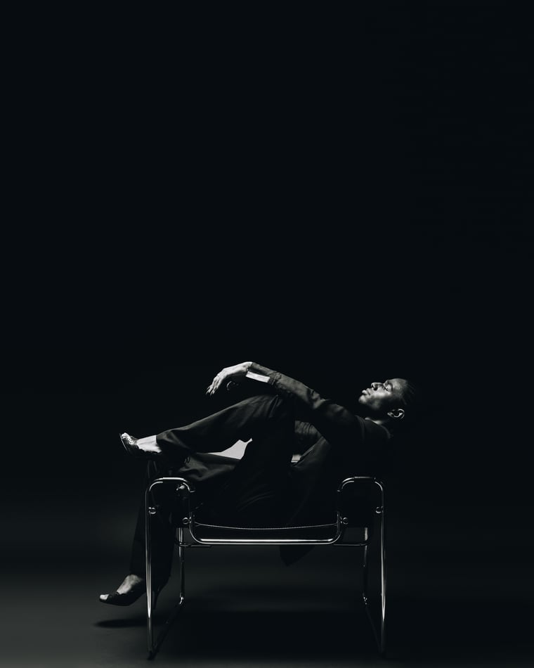A man in a black suit thrown back across a tubular steel sling chair in a small pool of light