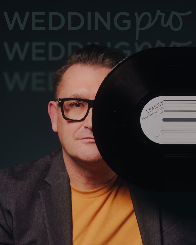 A man in heavy black glasses and an orange tee holding a vinyl record up beside his face