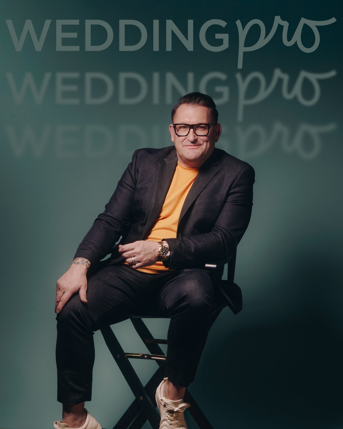 The same man seated on a director's chair with his legs crossed, against a teal wall of repeated brand lettering