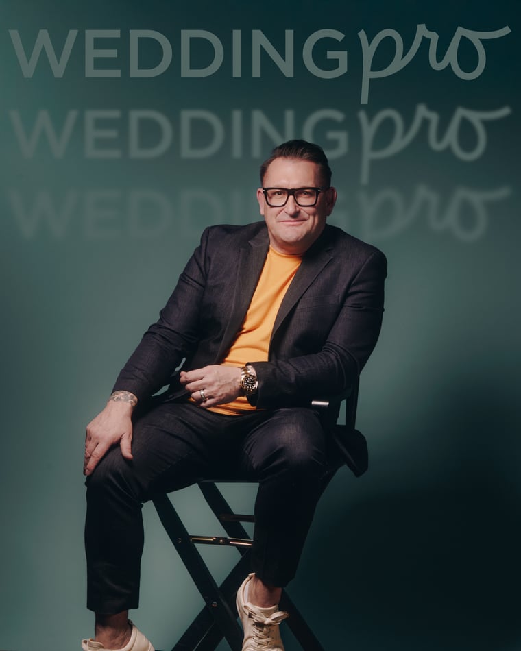 The same man seated on a director's chair with his legs crossed, against a teal wall of repeated brand lettering