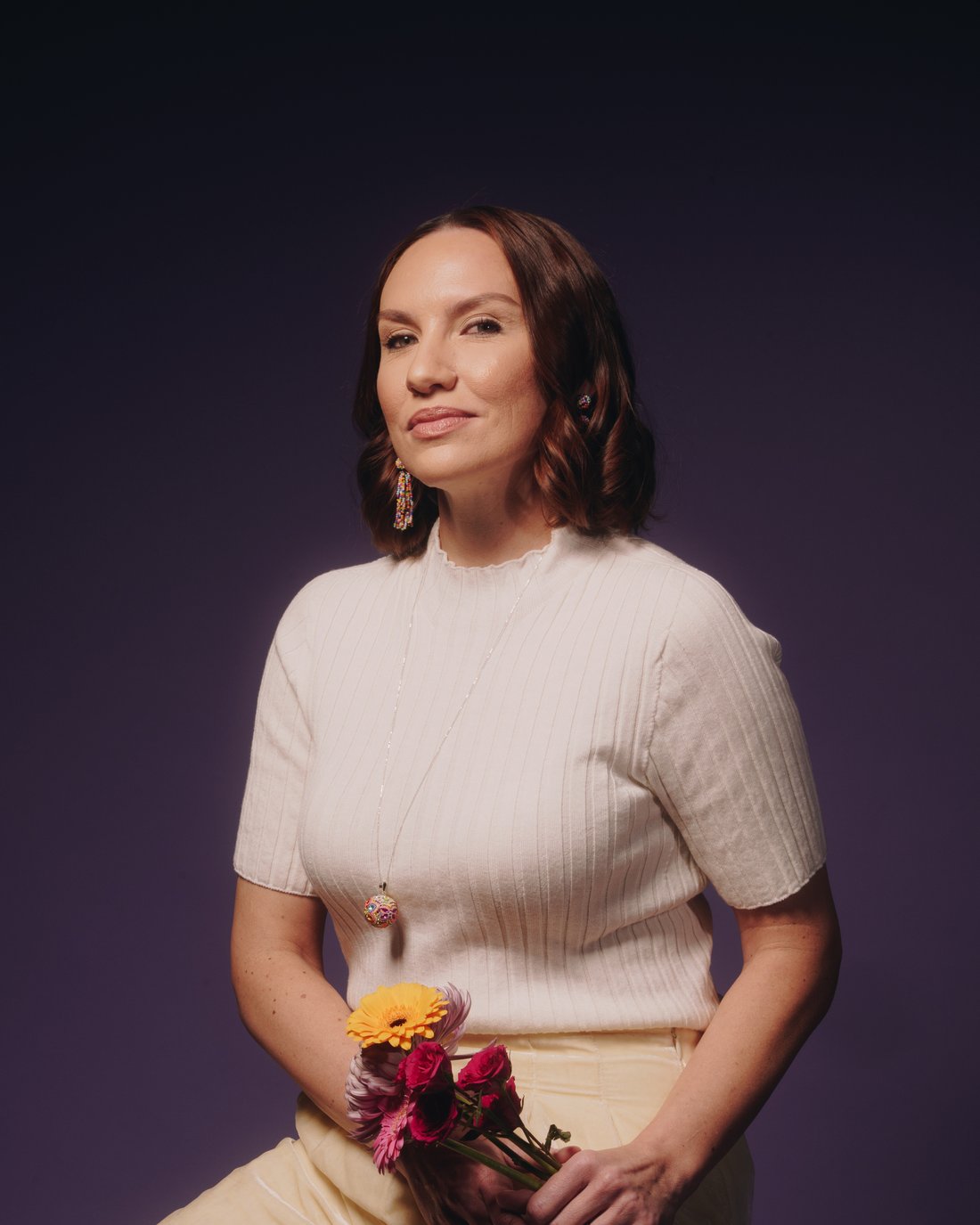 A woman in a cream ribbed knit holding a small bunch of orange and pink flowers against violet