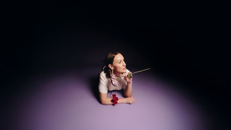 A woman lying on her front in a pool of lilac light, chin resting on her hands, a red bloom pinned at her collar