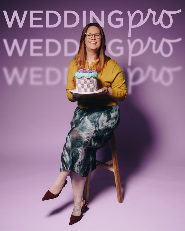 A woman in a mustard cardigan and painted silk skirt perched on a stool, holding out a chequerboard cake on a tray
