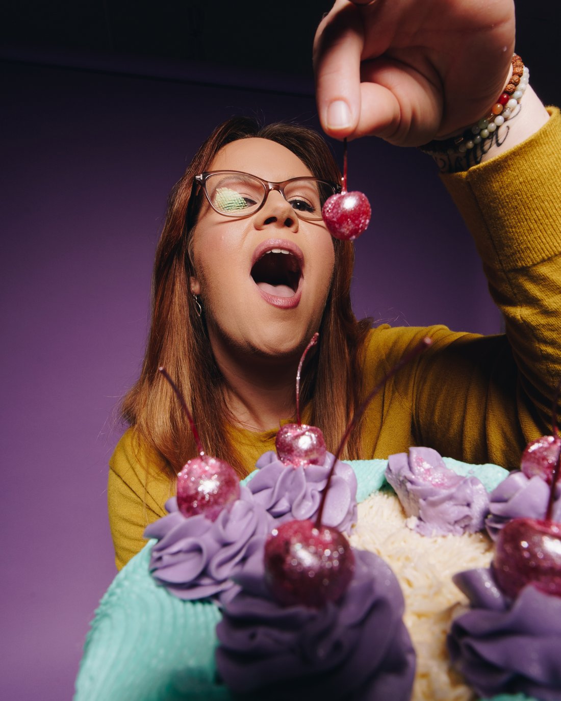 A woman in mustard yellow with her mouth wide open beneath a glittered cherry dangled above it, a purple ruffled cake in her other hand