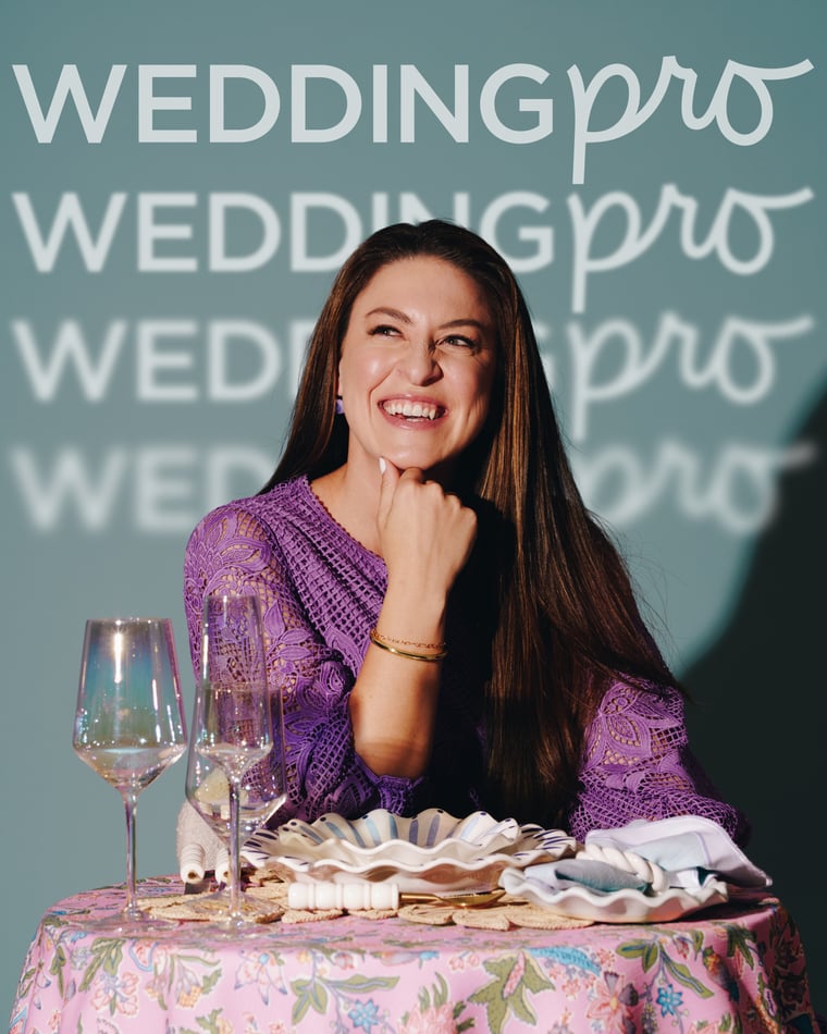 A woman in lilac lace laughing with her chin on her fist at a table set with wine glasses and scalloped china
