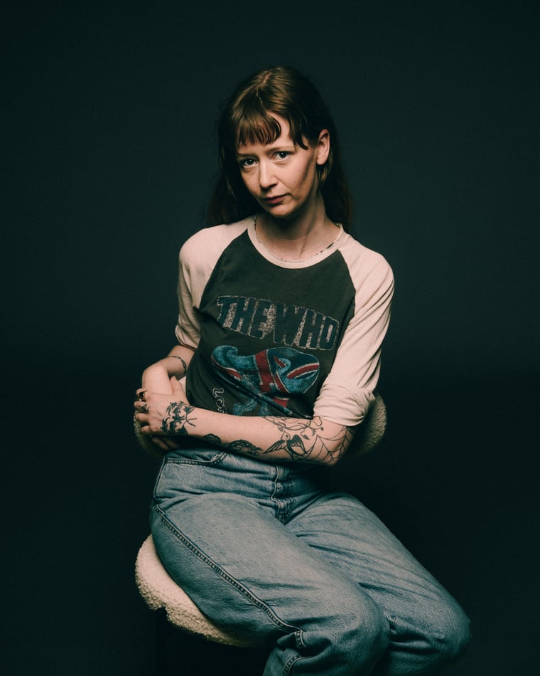 A woman with a blunt fringe in a worn band tee and baggy jeans, tattooed arms folded, perched on a low stool