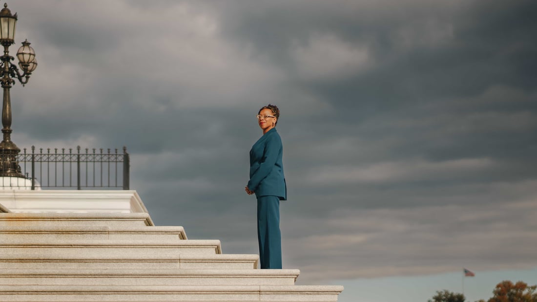 A woman in a teal suit standing alone at the top of a wide flight of marble steps under heavy grey cloud, an ornate cast iron lamppost at the edge of the frame
