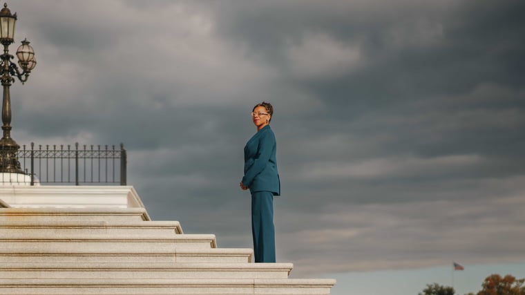 A woman in a teal suit standing alone at the top of a wide flight of marble steps under heavy grey cloud, an ornate cast iron lamppost at the edge of the frame