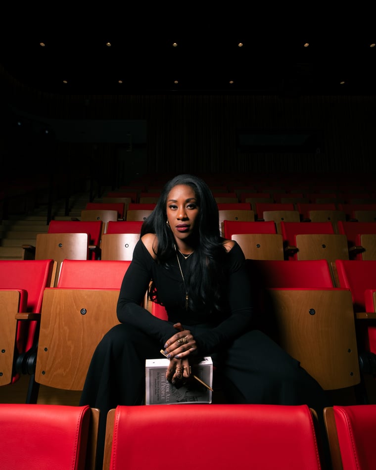 A woman in black sitting alone in a row of red theatre seats in an empty auditorium, a book in her lap