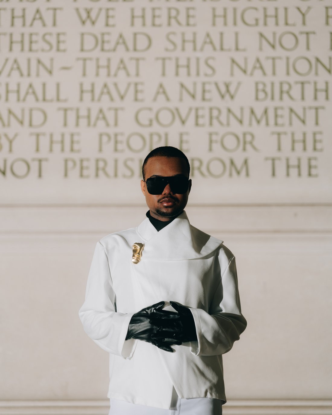 A figure in a sculptural white jacket and one long black glove, standing against a stone wall carved with the closing lines of the Gettysburg Address