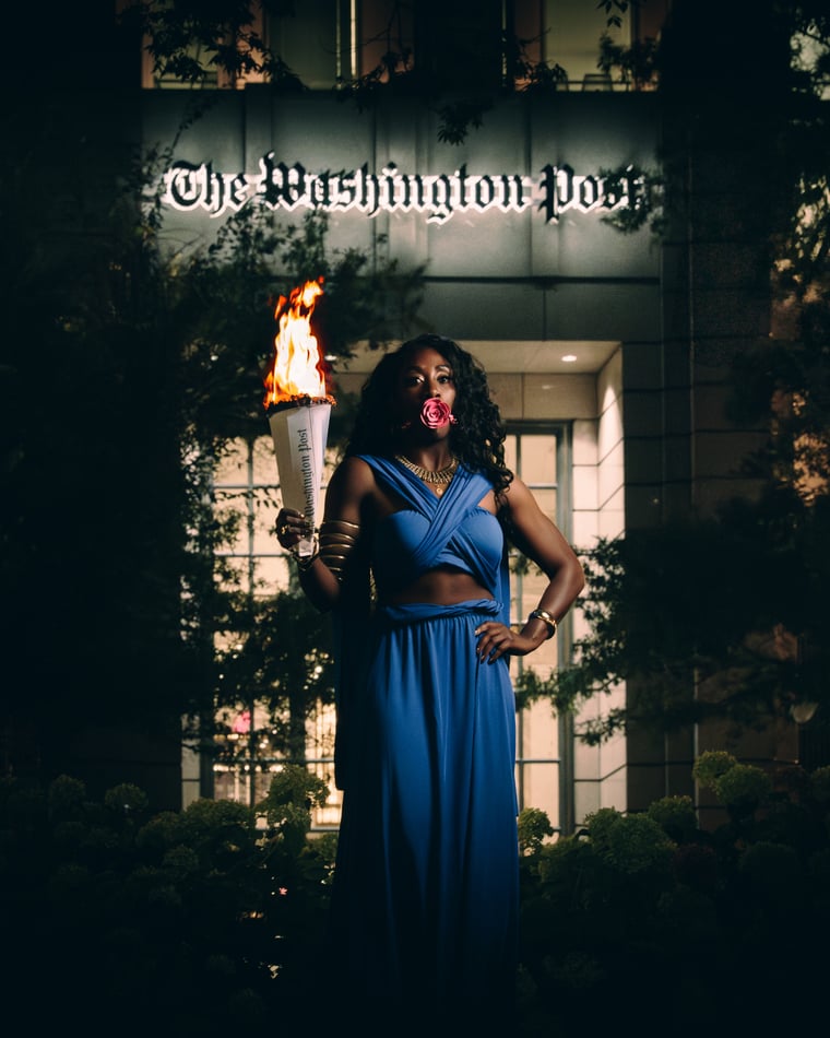 A woman in a draped blue gown holding a burning torch outside The Washington Post building at dusk