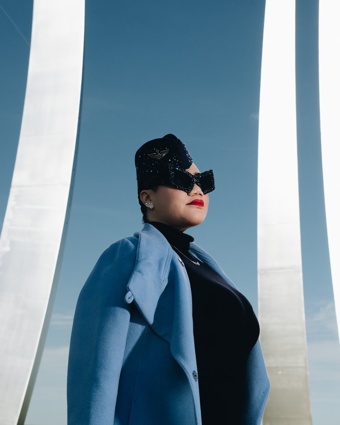 A woman in a pale blue coat, jewelled cap and oversized black sunglasses, chin lifted, standing between the white steel spires of the Air Force Memorial