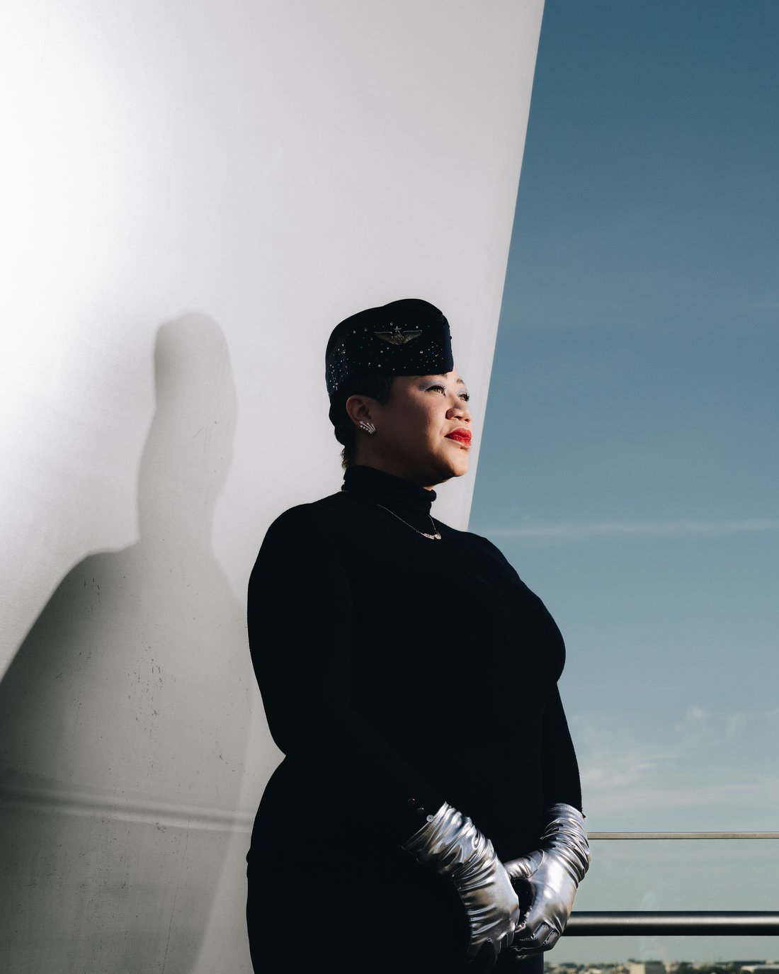 The same woman in black with silver opera gloves, in profile against a white curved wall that throws her shadow twice her size