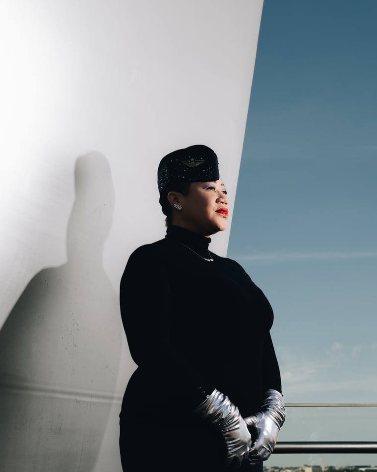 The same woman in black with silver opera gloves, in profile against a white curved wall that throws her shadow twice her size