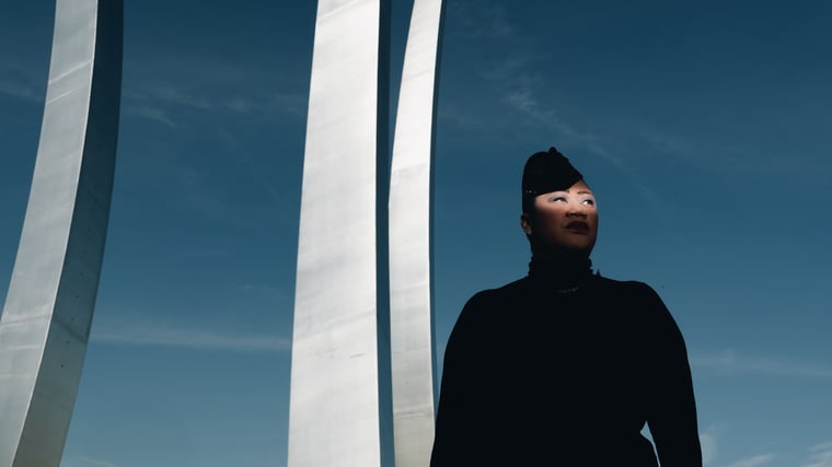 A near silhouette in a black turtleneck and beaded cap looking up, framed by three soaring white spires against open blue sky