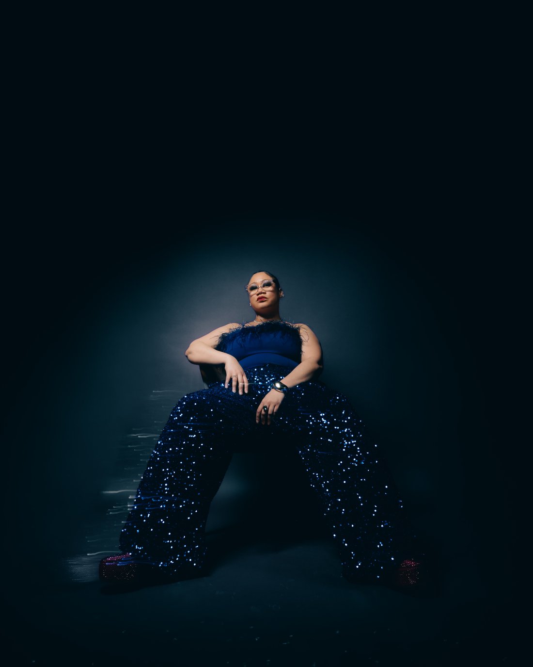 A woman in a navy sequinned gown sitting low on the floor, legs wide, held in a tight circle of light