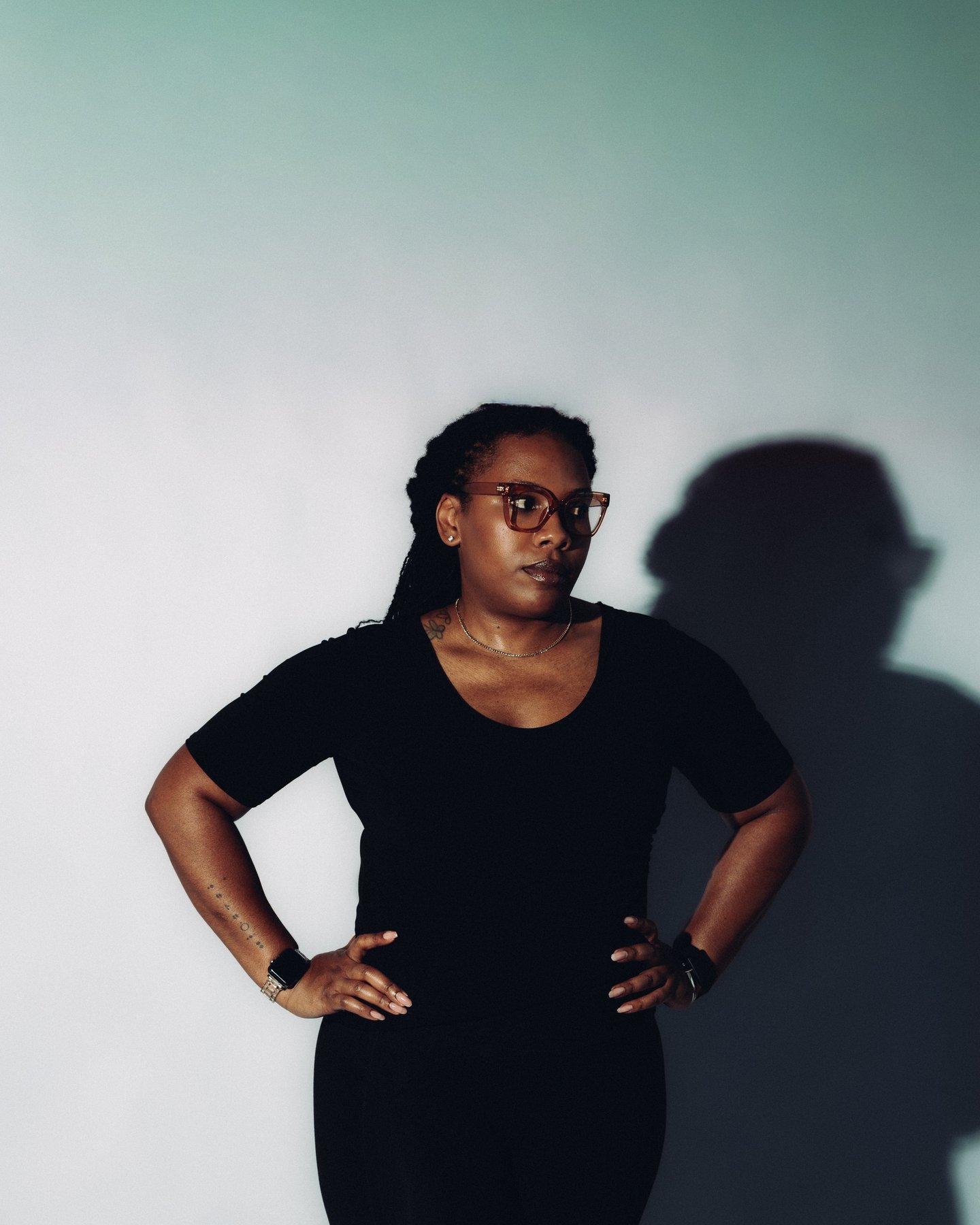 Standing in a black tee and glasses, a hand on the hip, a hard shadow on the wall