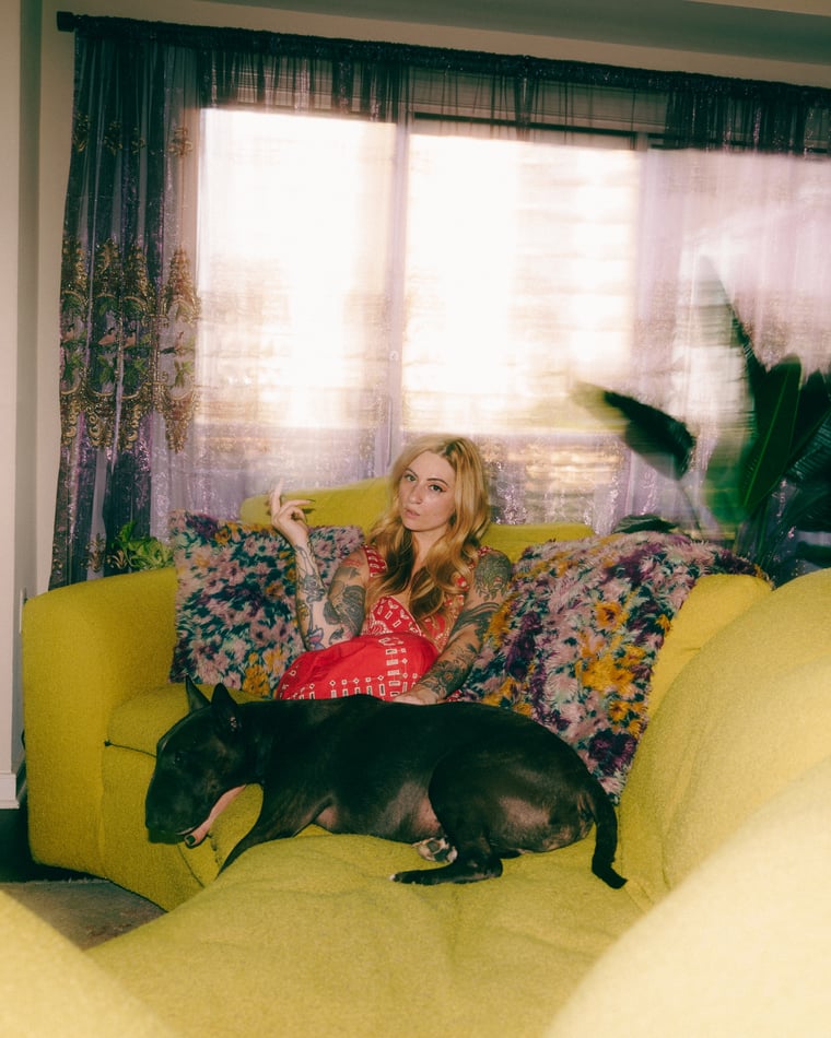 A woman on a yellow sofa with a black French bulldog asleep beside her, late light coming through the curtains
