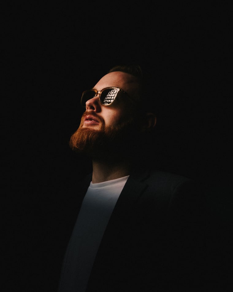 A bearded man in mirrored sunglasses tilting his face up into a single shaft of light