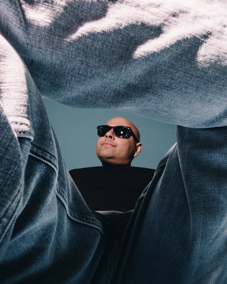 Shot from the ground between a pair of denim clad legs, a man in dark glasses looks down into the lens against blue sky
