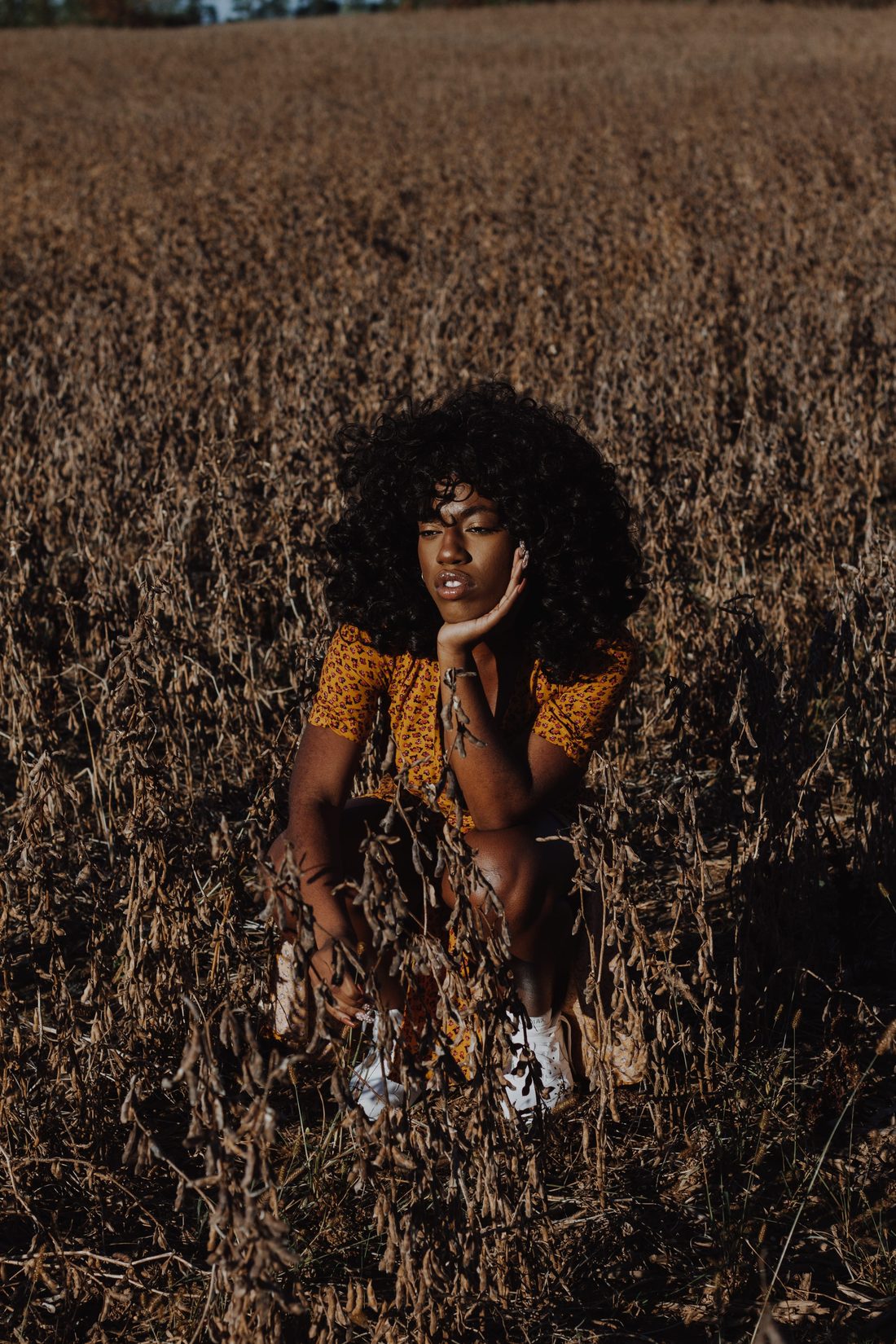A woman with an afro crouched low in a harvested field, chin resting on her hand