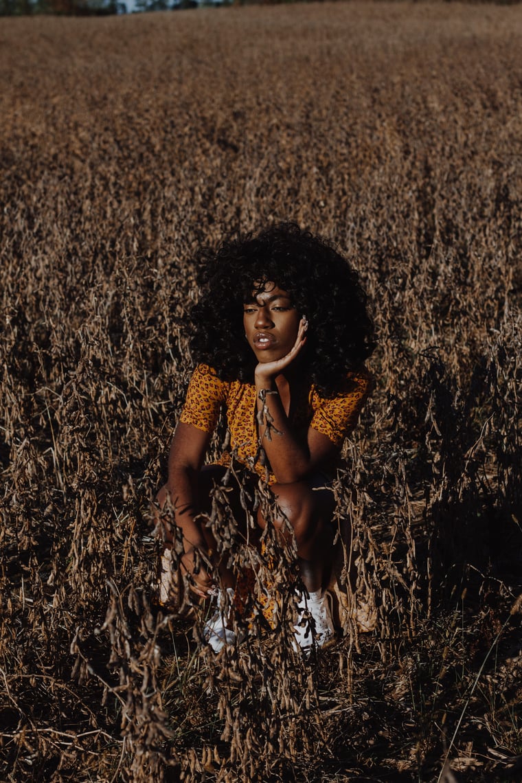 A woman with an afro crouched low in a harvested field, chin resting on her hand