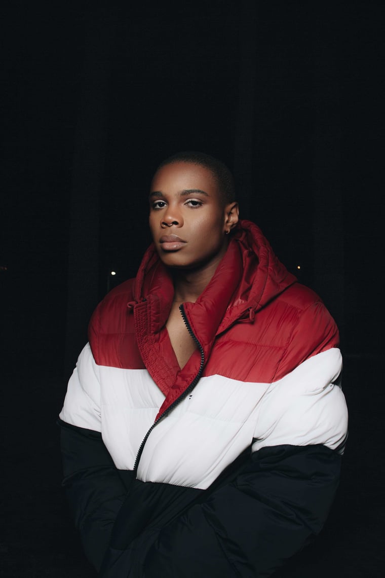 A person in a red, white and black colour blocked puffer jacket, arms folded, against black