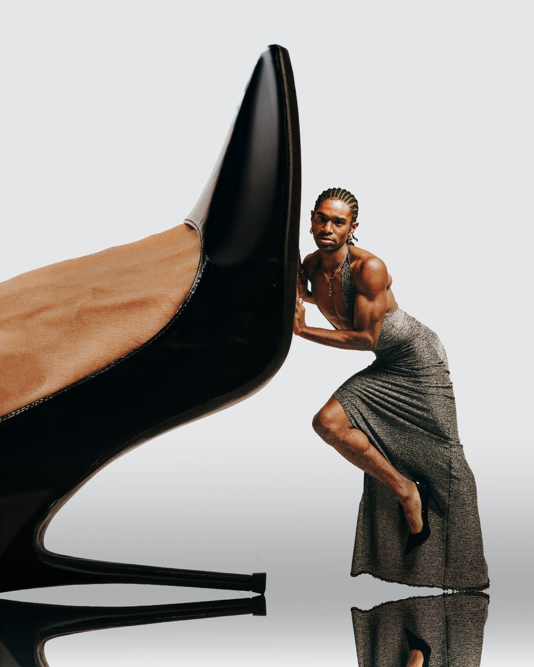 A dancer in a grey draped dress braced against an enormous black stiletto on a white sweep, dwarfed by it